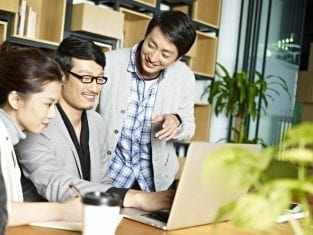 Orang-orang bekerja bersama di depan laptop; satu orang berdiri menunjuk layar, lainnya duduk memperhatikan, di dalam kantor dengan rak buku dan tanaman.
